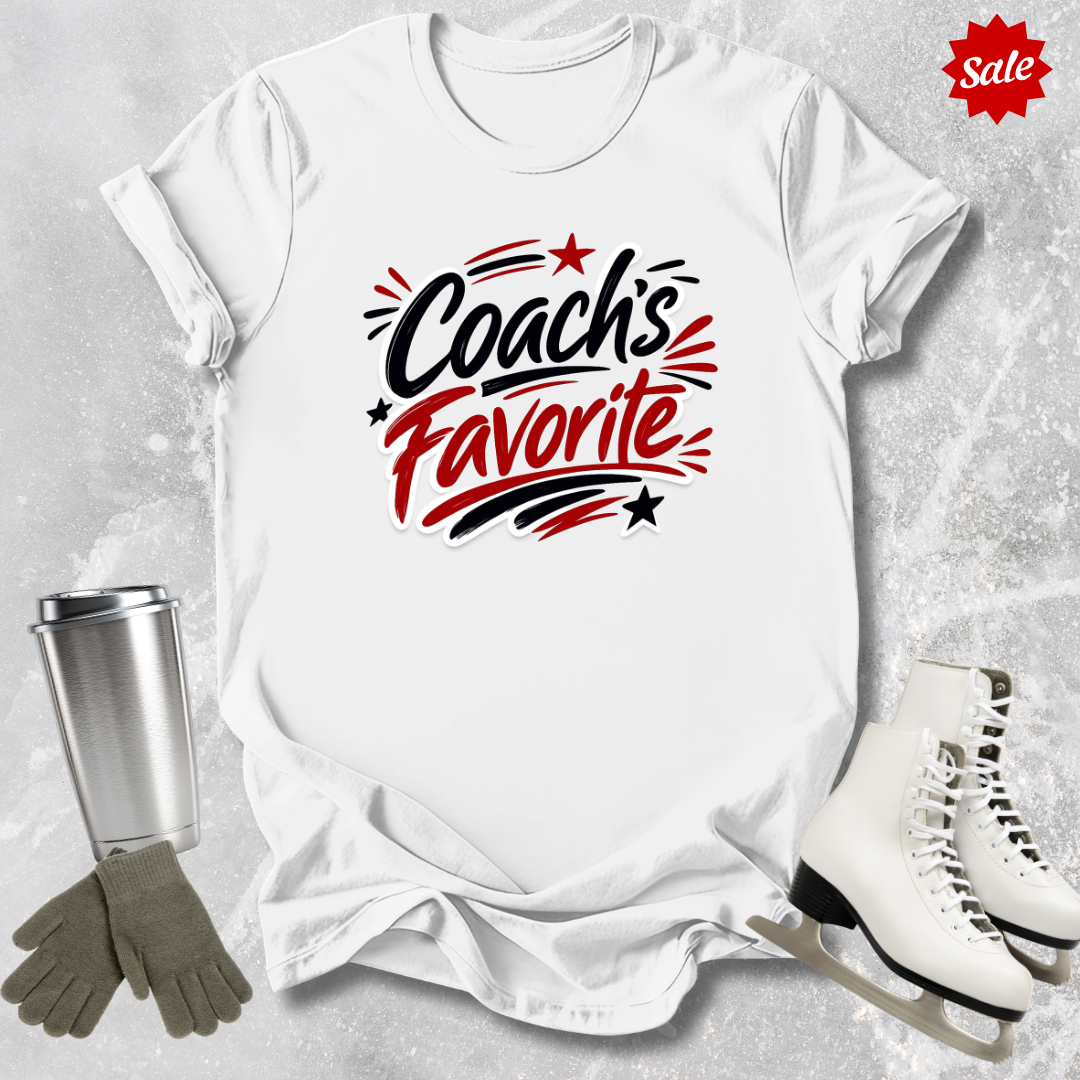 White t-shirt with 'Coach's Favorite' text  for figure skaters, surrounded by a silver tumbler, gray gloves, and white skates on a textured surface.
