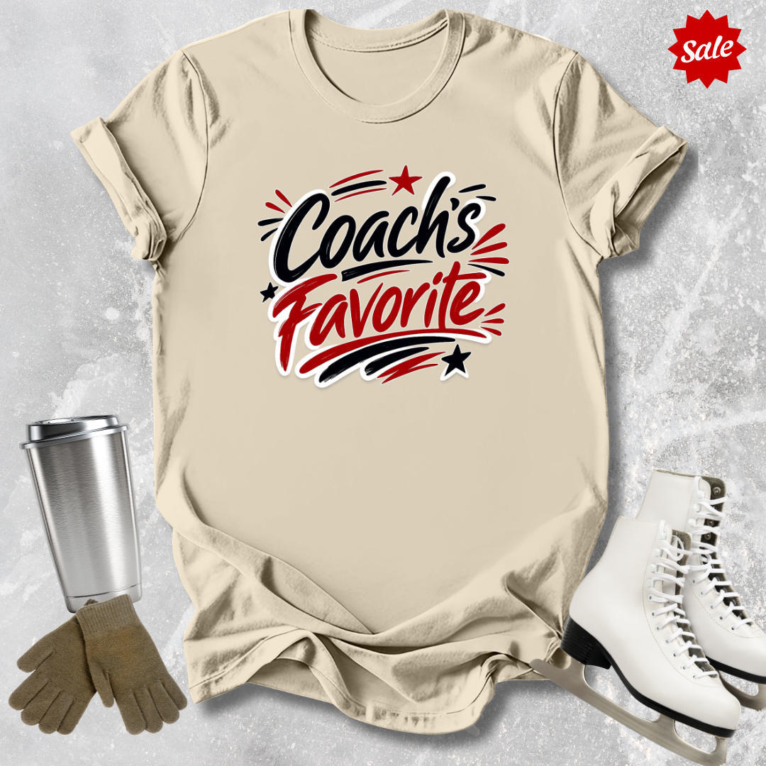 Beige t-shirt with 'Coach's Favorite' text  for figure skaters, silver tumbler, brown gloves, and white sneakers on a textured gray background.