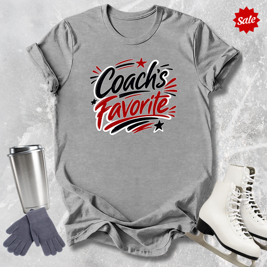 Gray t-shirt with 'Coach's Favorite' text   for figure skaters, surrounded by a silver tumbler, gray gloves, and white skates on a textured surface.