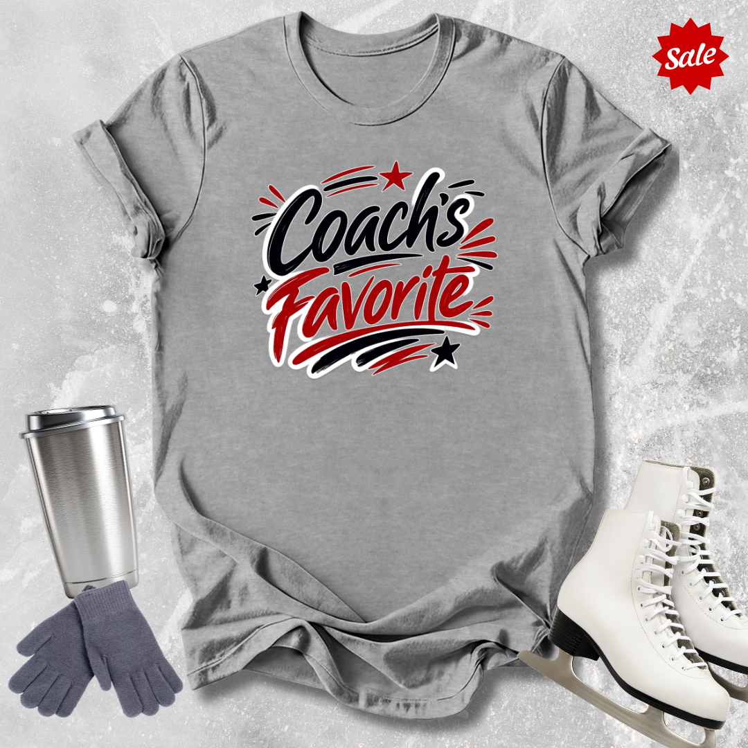 Gray t-shirt with 'Coach's Favorite' text  for figure skaters, surrounded by a silver tumbler, gray gloves, and white skates on a textured surface.