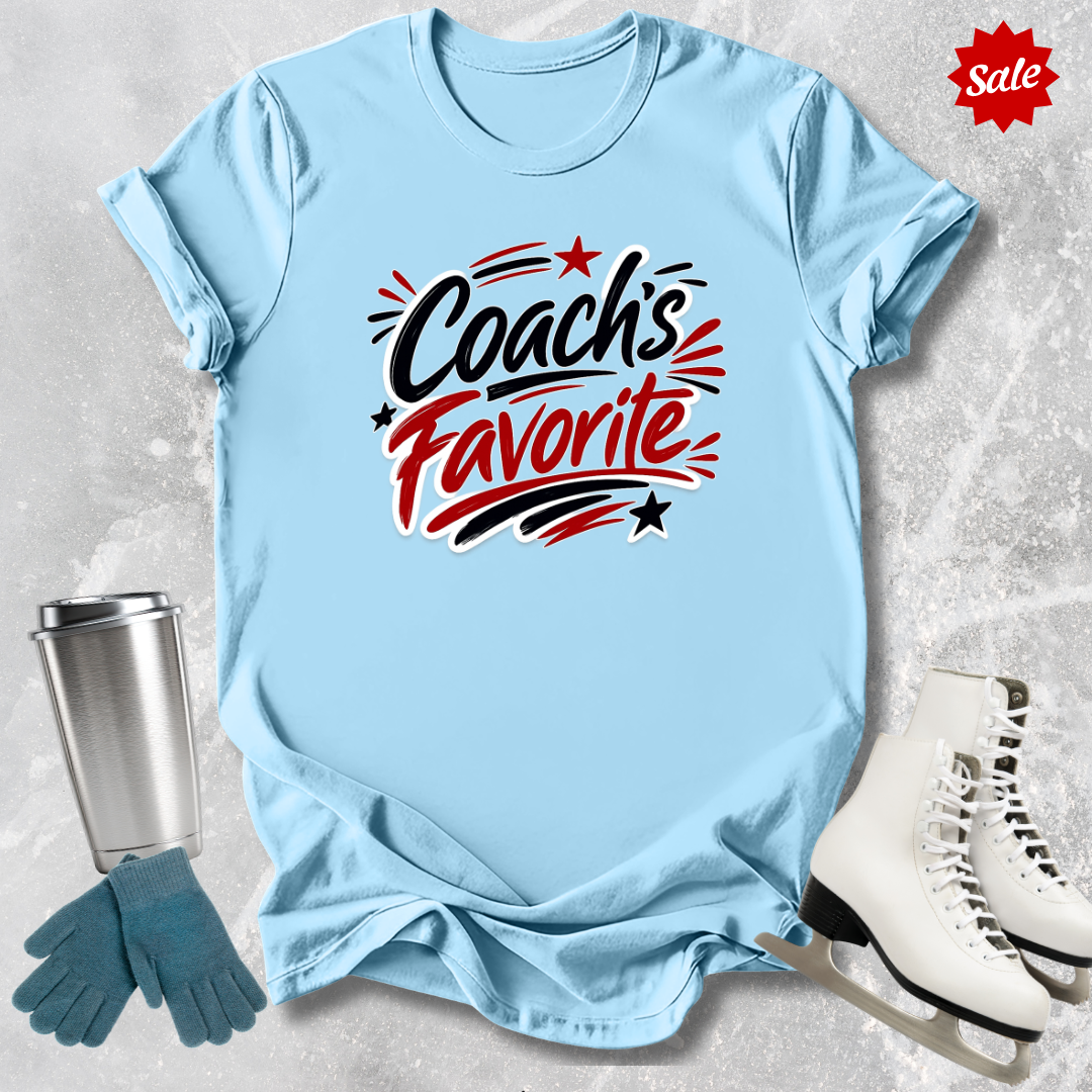 Light blue t-shirt with 'Coach's Favorite' text  for figure skaters, next to a silver tumbler, green gloves, and white skates on a textured gray background.
