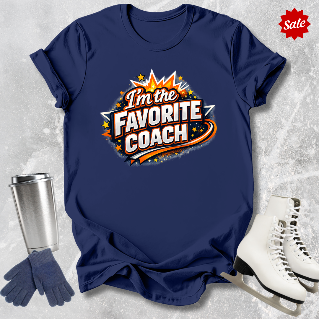 Navy blue t-shirt with 'I'm the Favorite Coach' design  for figure skating coaches, next to a silver tumbler, gloves, and ice skates on a textured gray background.