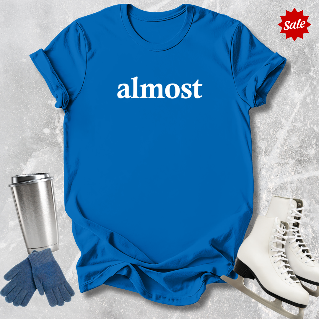 Blue t-shirt with 'almost' text for figure skating coaches,, silver tumbler, blue gloves, and white skates on a textured gray background.