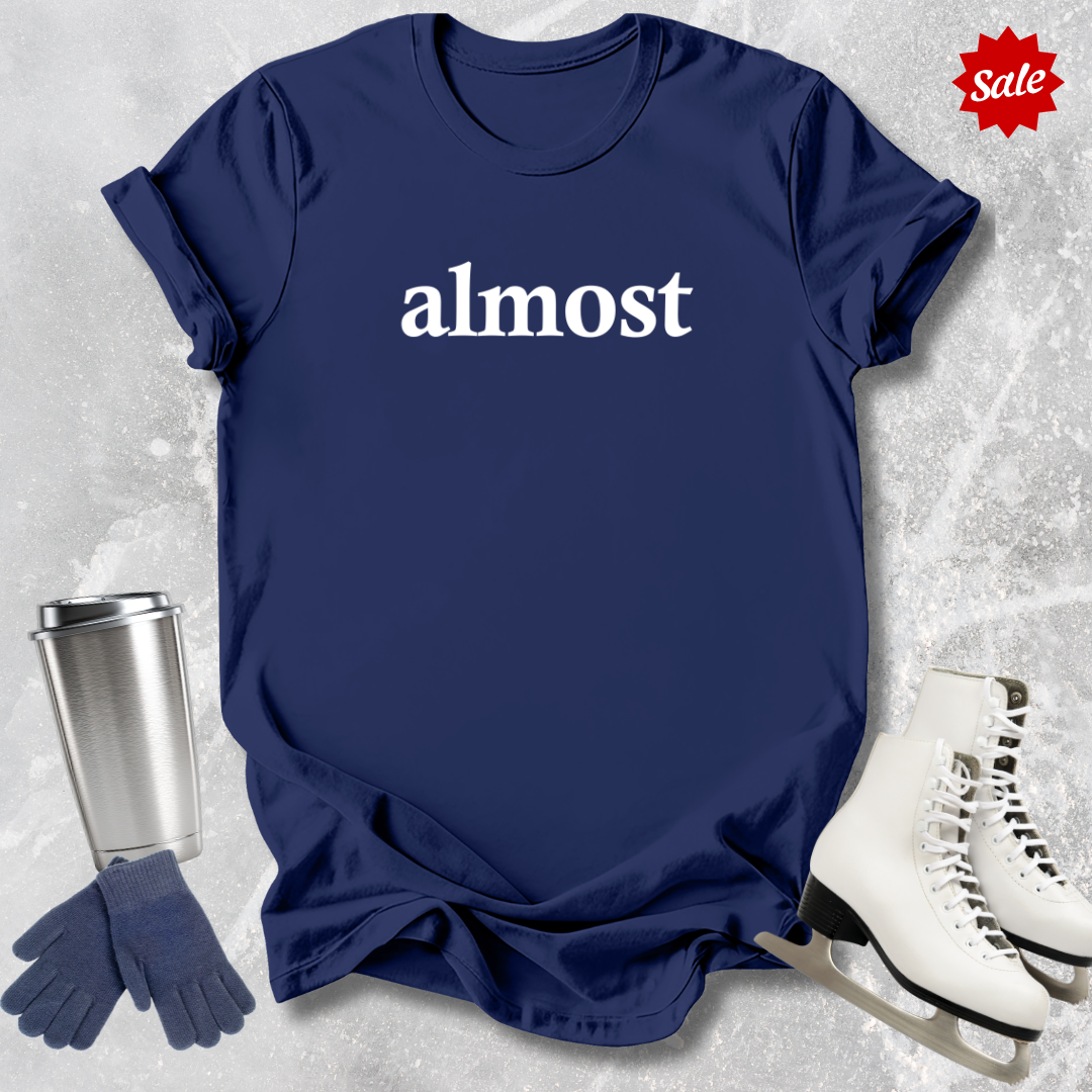 Navy blue t-shirt with 'almost' text  for figure skating coaches,, silver tumbler, gloves, and ice skates on a textured gray background.