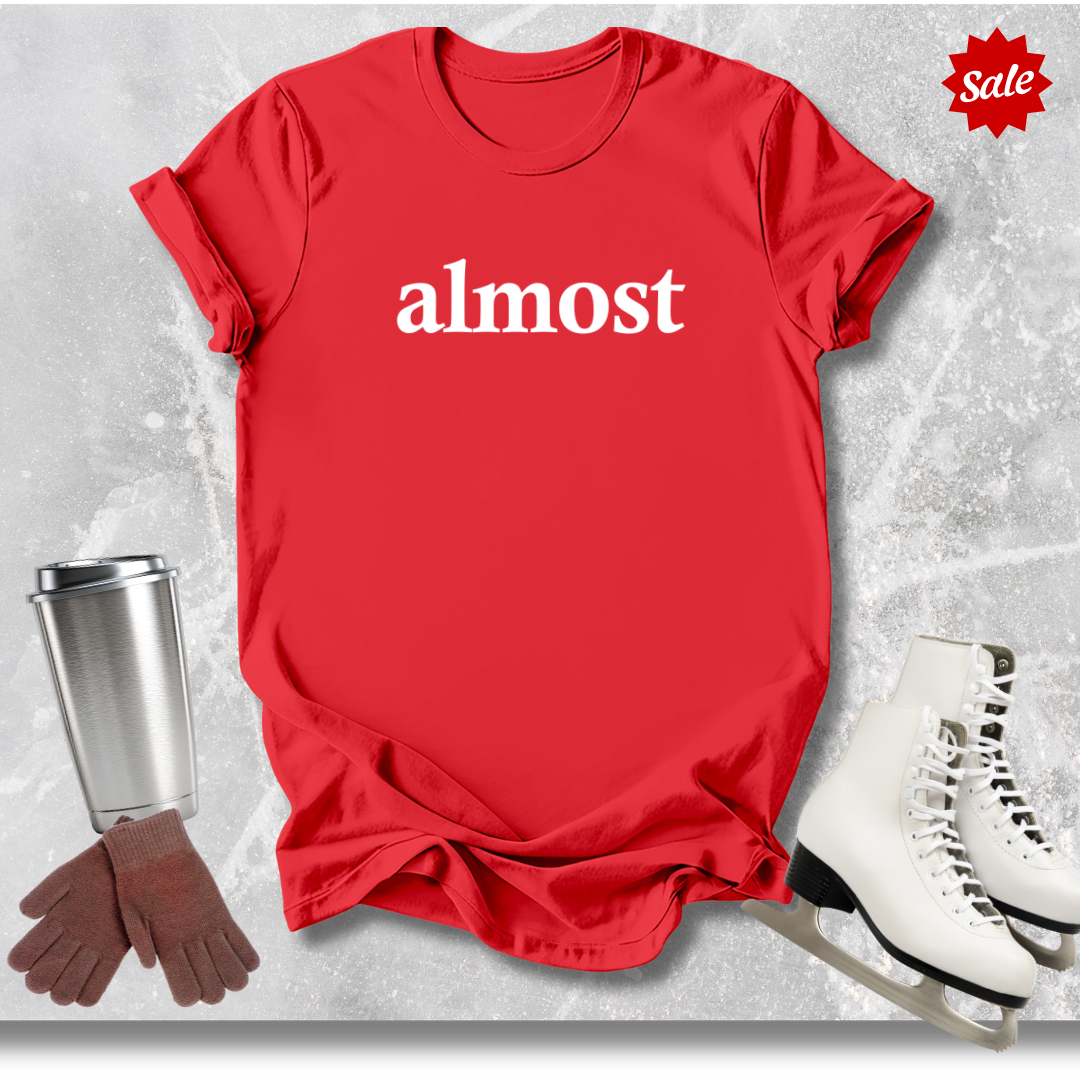 Red t-shirt with 'almost' text for figure skating coaches, white skates, silver tumbler, and brown gloves on a textured gray background.