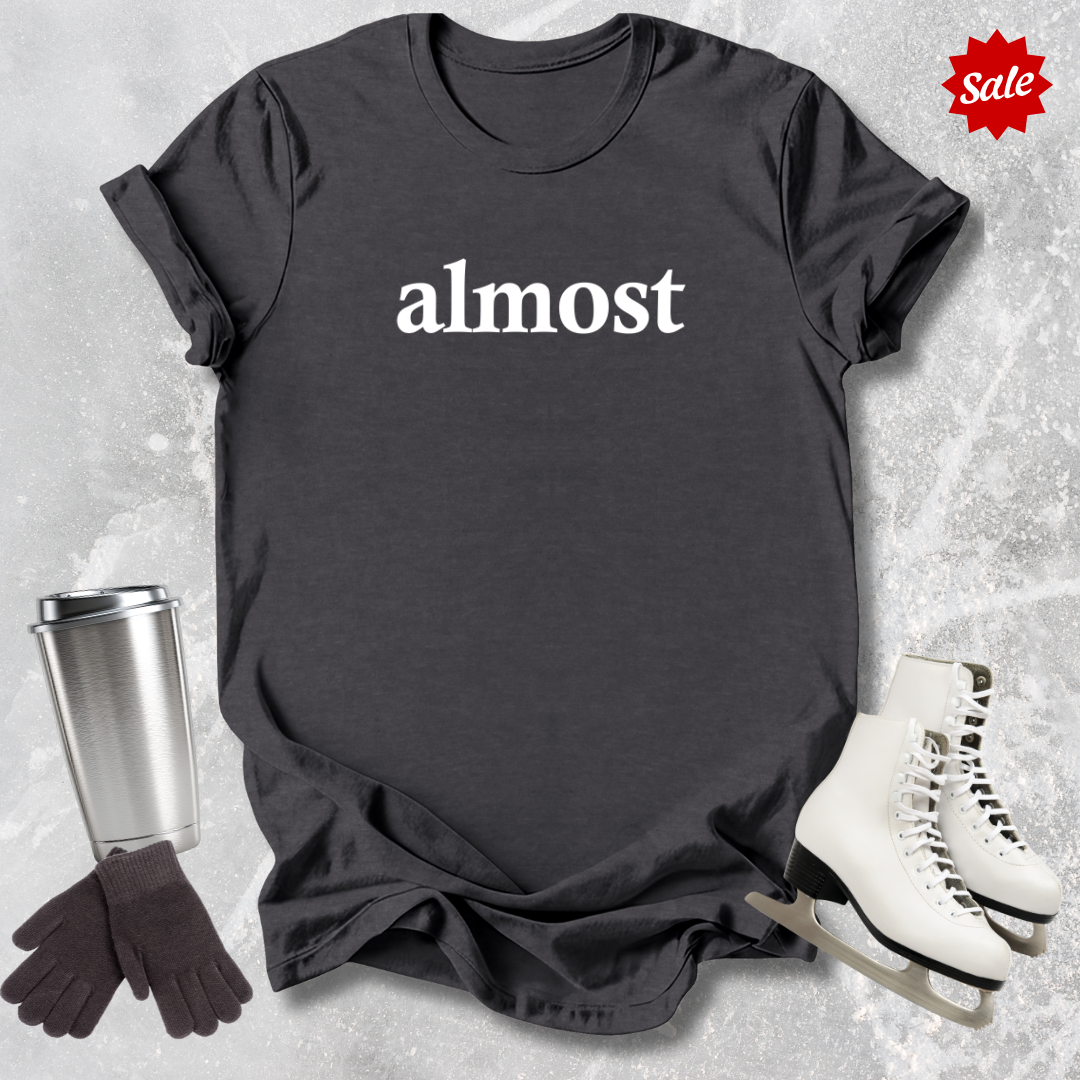 Black t-shirt with 'almost' text for figure skating coaches,, silver tumbler, black gloves, white skates on a textured surface.