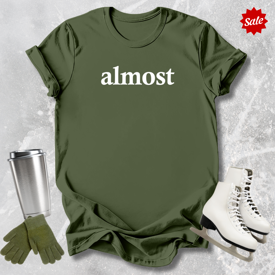 Green t-shirt with 'almost' text  for figure skating coaches,, silver tumbler, green gloves, and white skates on a textured gray background.