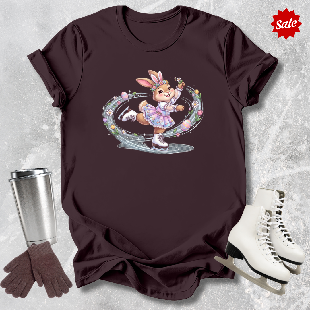 Brown t-shirt with a cartoon a figure skating Easter bunny design, next to a silver tumbler, brown gloves, and white ice skates on a textured gray background.