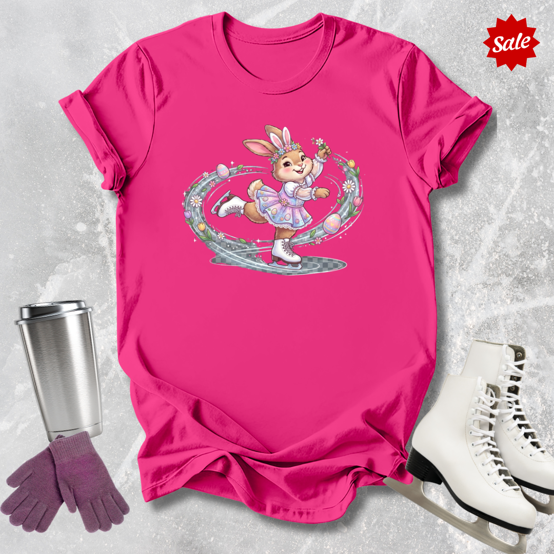 Pink t-shirt with a cartoon character a figure skating Easter bunny graphic, next to a silver tumbler, purple gloves, and white ice skates on a light gray background.