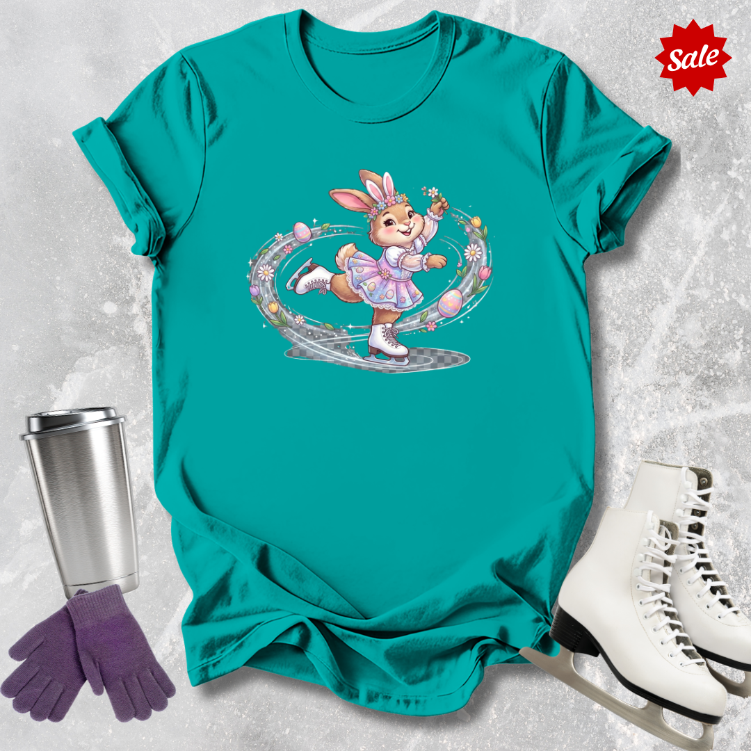 Teal t-shirt with a cartoon a figure skating Easter bunny design, next to a silver tumbler, purple gloves, and white skates on a textured gray background.