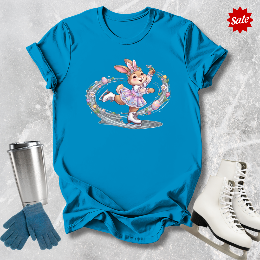 Blue t-shirt with a cartoon a figure skating Easter bunny design, next to a silver tumbler, blue gloves, and white ice skates on a textured gray background.