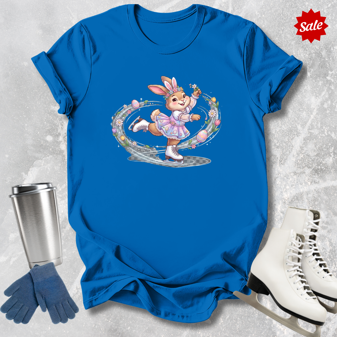 Blue t-shirt with a cartoon a figure skating Easter bunny design, next to a silver tumbler, blue gloves, and white ice skates on a textured gray background.