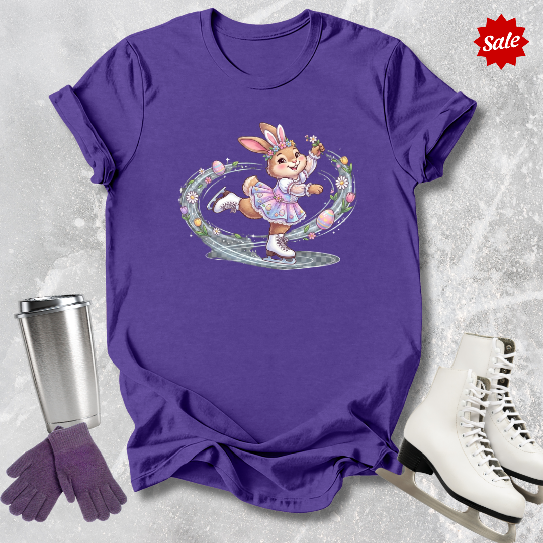 Purple t-shirt with a cartoon a figure skating Easter bunny design, silver tumbler, purple gloves, and white ice skates on a textured gray background.