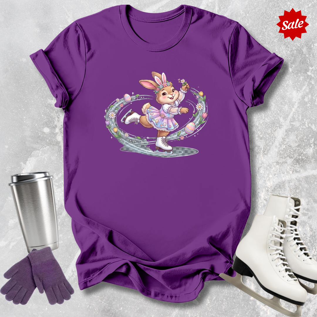 Purple t-shirt with a figure skating Easter bunny graphic, next to a silver tumbler, purple gloves, and white skates on a light gray background.