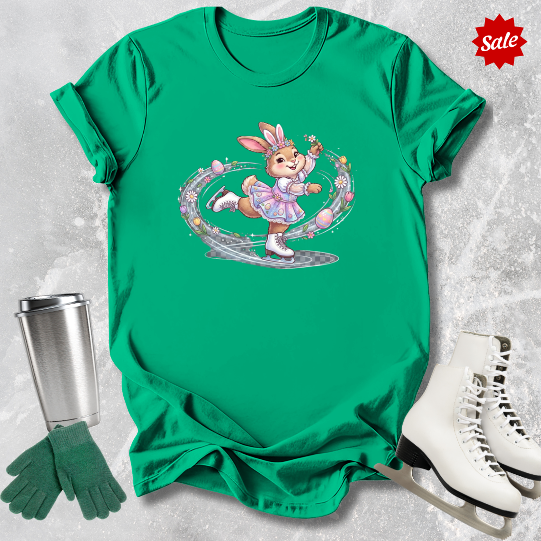 Green t-shirt with a cartoon a figure skating Easter bunny design, next to a silver tumbler, green gloves, and white skates on a light gray background.
