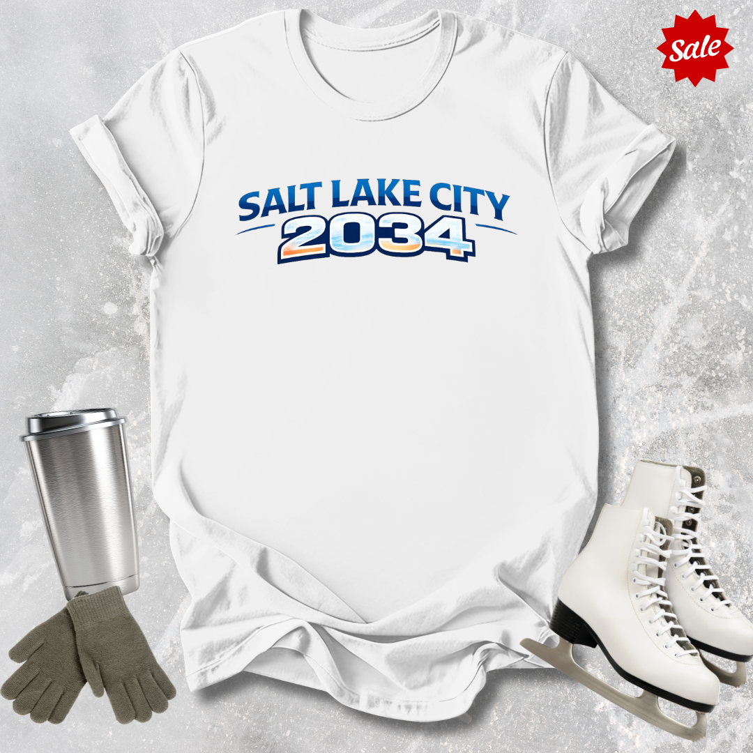 White t-shirt with 'Salt Lake City 2034' text, surrounded by ice skates, gloves, and a tumbler on a textured surface.
