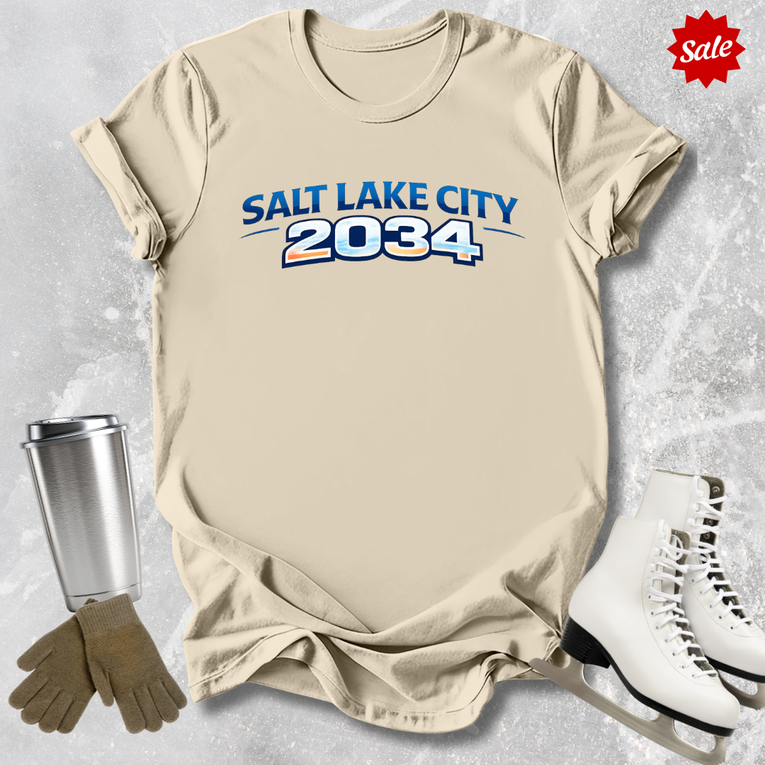 Beige t-shirt with 'Salt Lake City 2034' text, next to a silver tumbler, brown gloves, and white skates on a textured gray background.