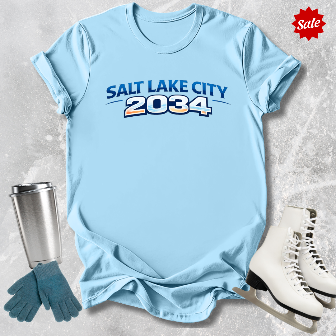Light blue t-shirt with 'Salt Lake City 2034' text, next to a silver tumbler, green gloves, and white skates on a light gray background.
