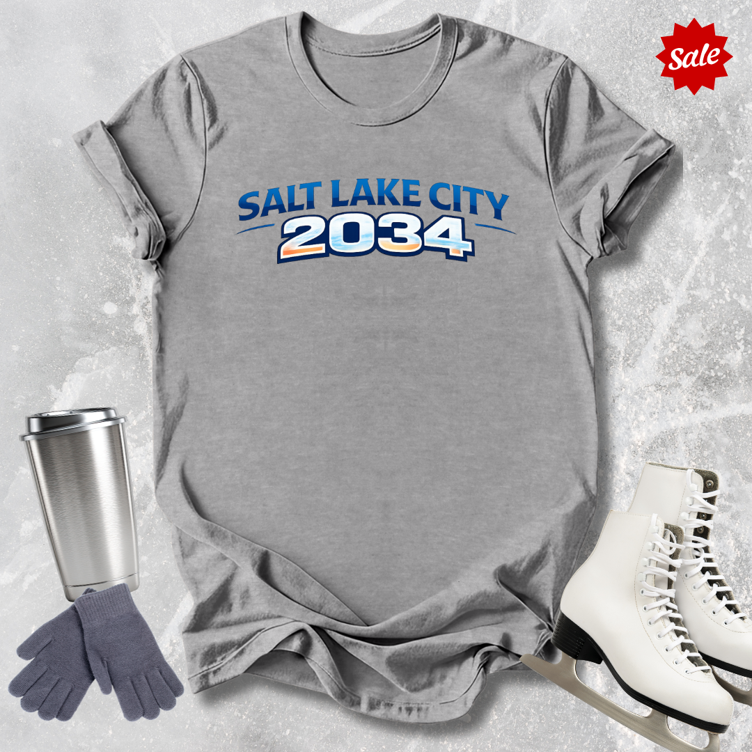 Gray t-shirt with 'Salt Lake City 2034' text, surrounded by a silver tumbler, gray gloves, and white skates on a light gray background.
