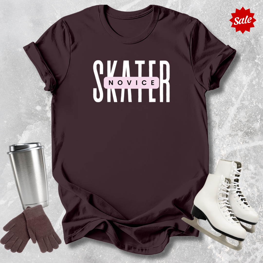 Maroon t-shirt with 'SKATER NOVICE' text, silver tumbler, brown gloves, and white skates on a textured gray background.