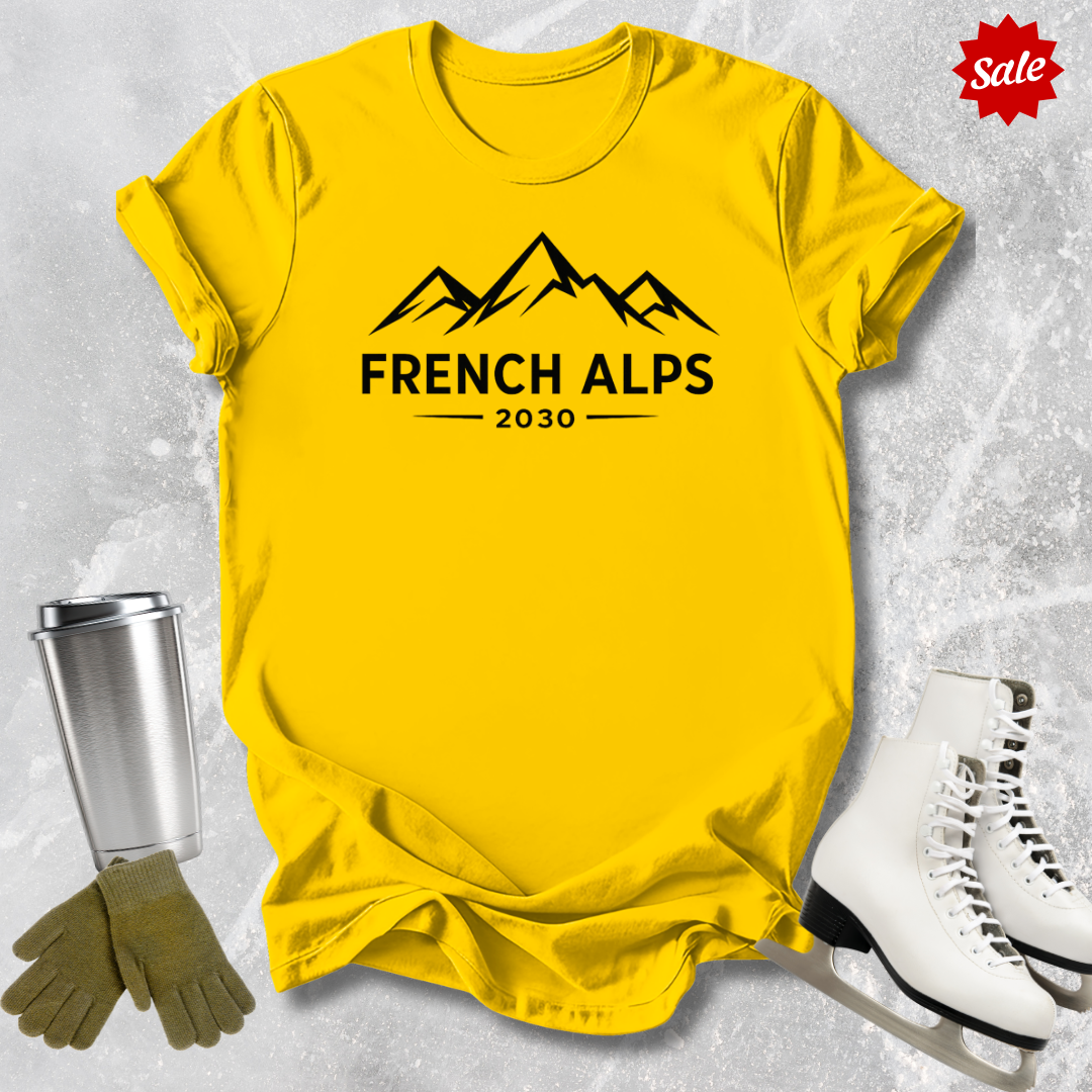 Yellow t-shirt with 'French Alps 2030' text, surrounded by winter accessories on a textured gray background.