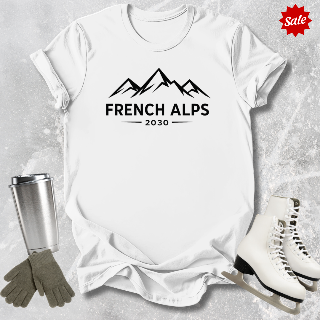 White t-shirt with 'French Alps 2030' design, surrounded by ice skates, gloves, and a tumbler on a textured gray background.