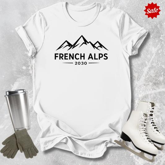 White t-shirt with 'French Alps 2030' design, surrounded by ice skates, gloves, and a tumbler on a textured gray background.