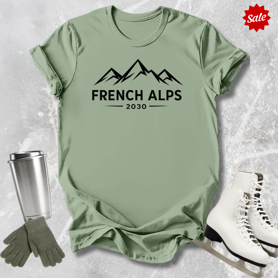 Green t-shirt with 'French Alps 2030' text and graphics, surrounded by winter accessories on a textured surface.