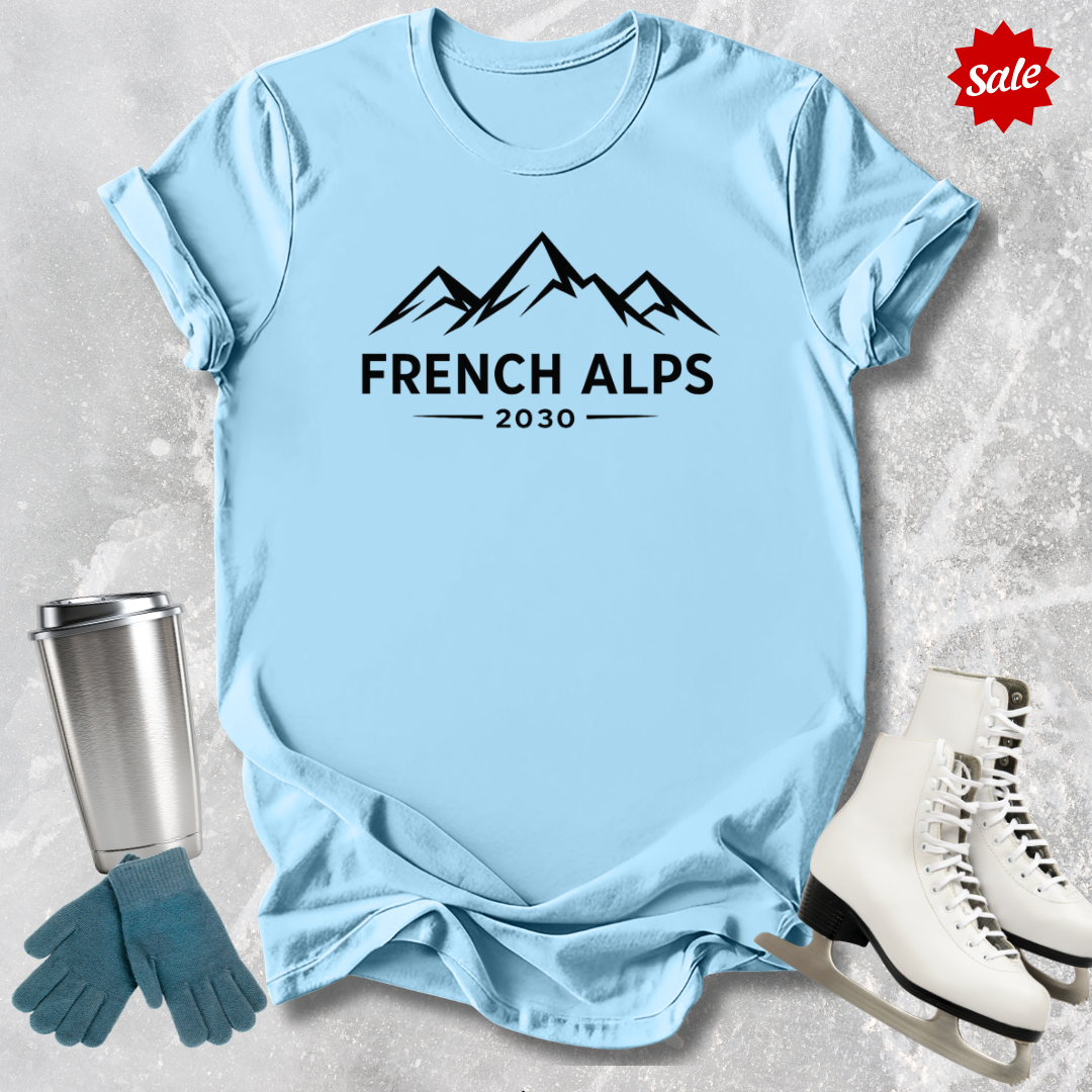 Light blue t-shirt with 'French Alps 2030' design, next to a silver tumbler, green gloves, and white skates on a textured surface.