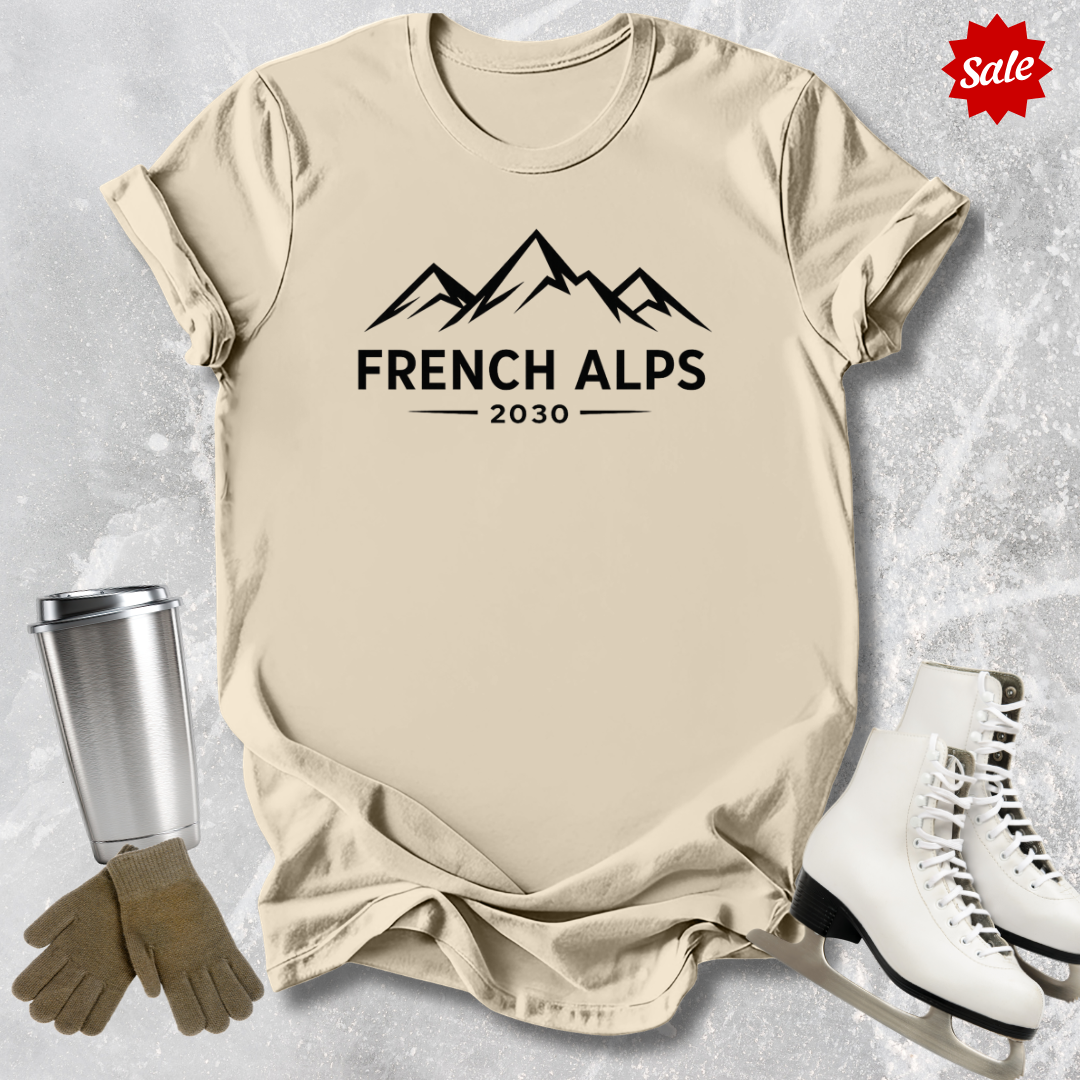 Beige t-shirt with 'French Alps 2030' text, silver tumbler, brown gloves, and white skates on a textured gray background.