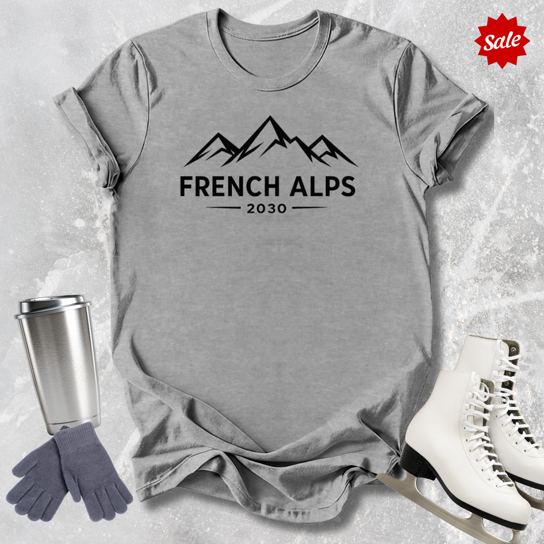 Gray t-shirt with 'French Alps 2030' text, silver tumbler, gray gloves, and white skates on a textured surface.