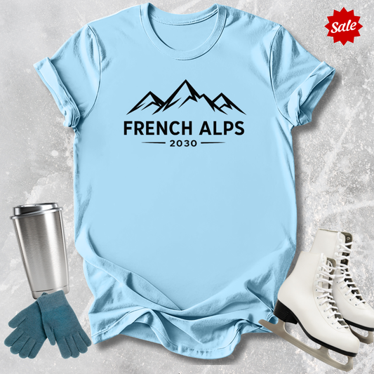 Light blue t-shirt with 'French Alps 2030' design, next to a silver tumbler, green gloves, and white skates on a textured surface.