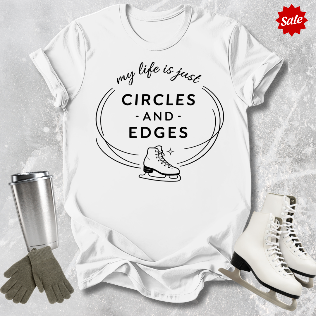 White t-shirt with 'my life is just circles and edges' text and ice skates, surrounded by ice skates and a tumbler on a textured surface.