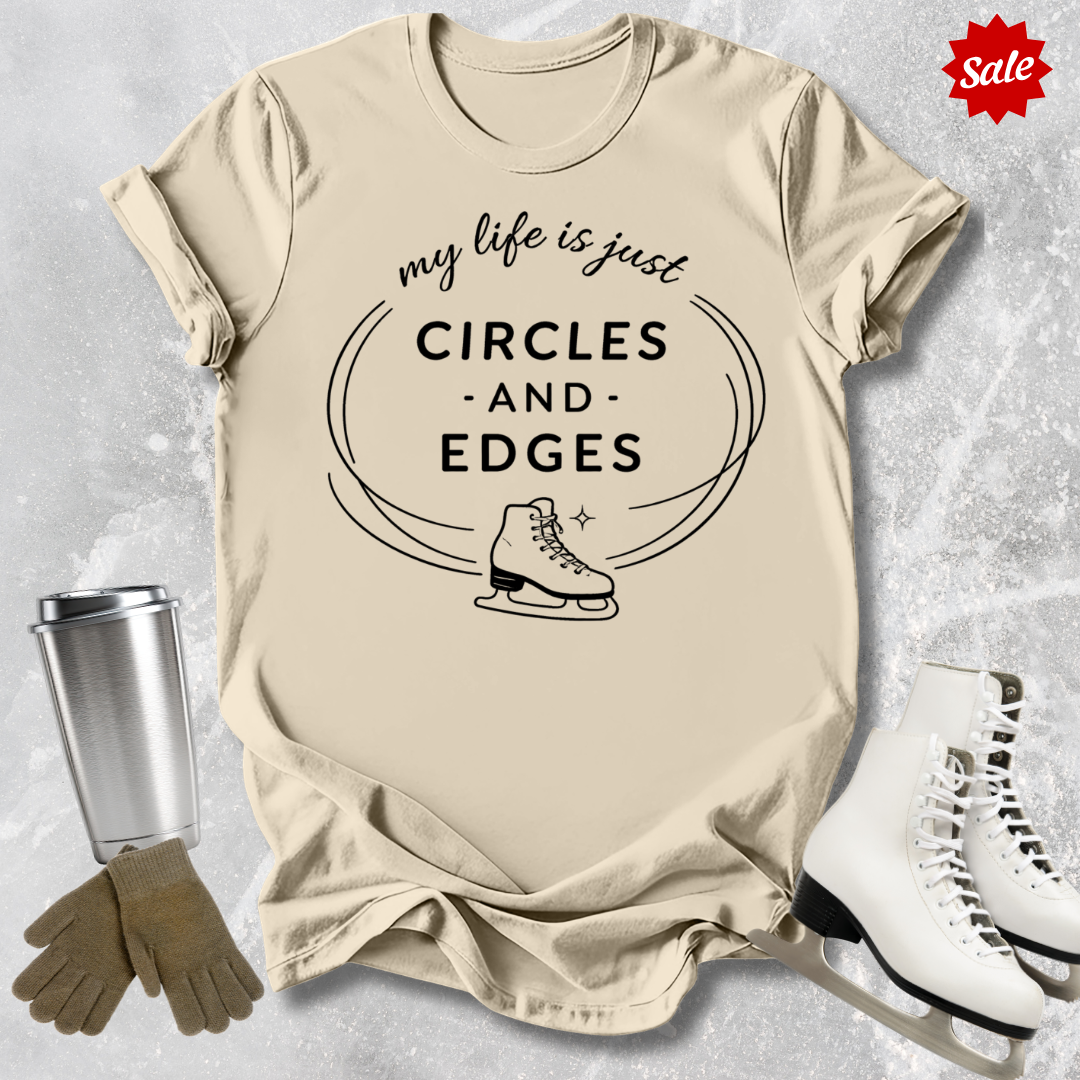 Beige t-shirt with 'my life is just circles and edges' text and ice skates, surrounded by ice skates and a tumbler on a textured surface.