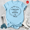 Light blue t-shirt with 'my life is just circles and edges' text and ice skate graphic, surrounded by ice skates and gloves on a textured surface.