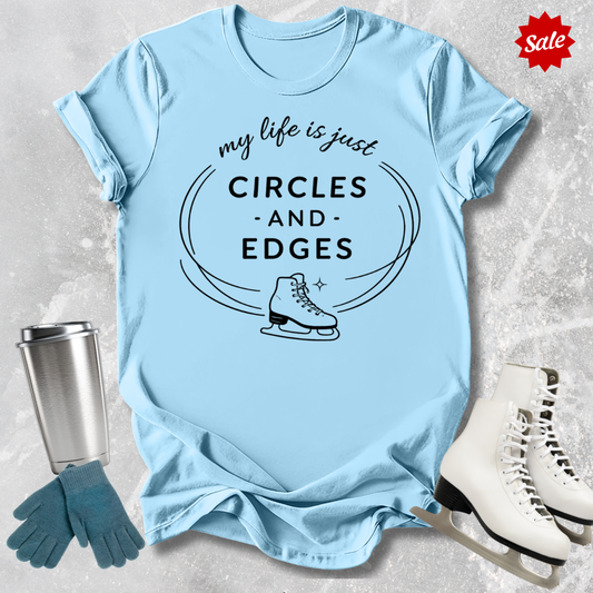 Light blue t-shirt with 'my life is just circles and edges' text and ice skate graphic, surrounded by ice skates and gloves on a textured surface.