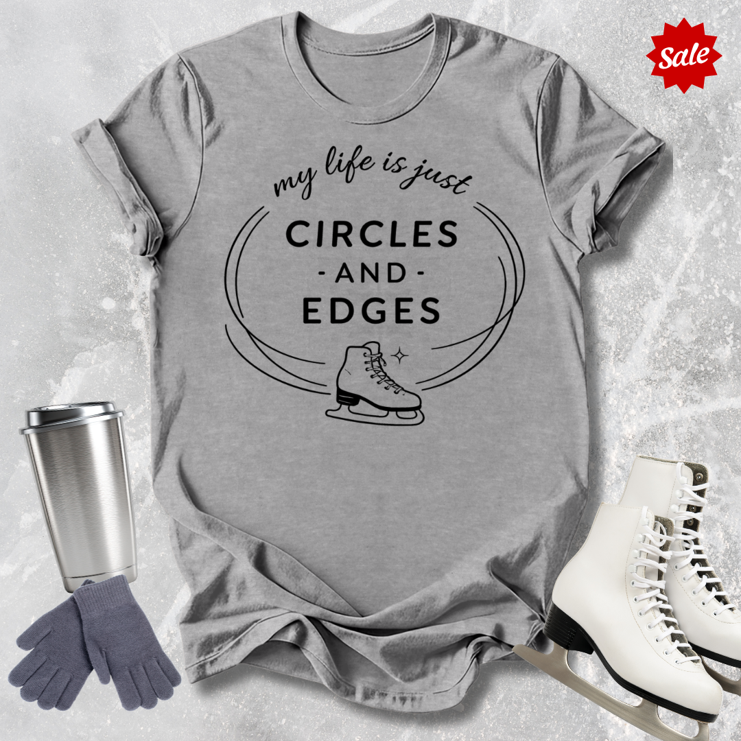 Gray  t-shirt with 'my life is just circles and edges' text and ice skates, surrounded by ice skates and a tumbler on a textured surface.