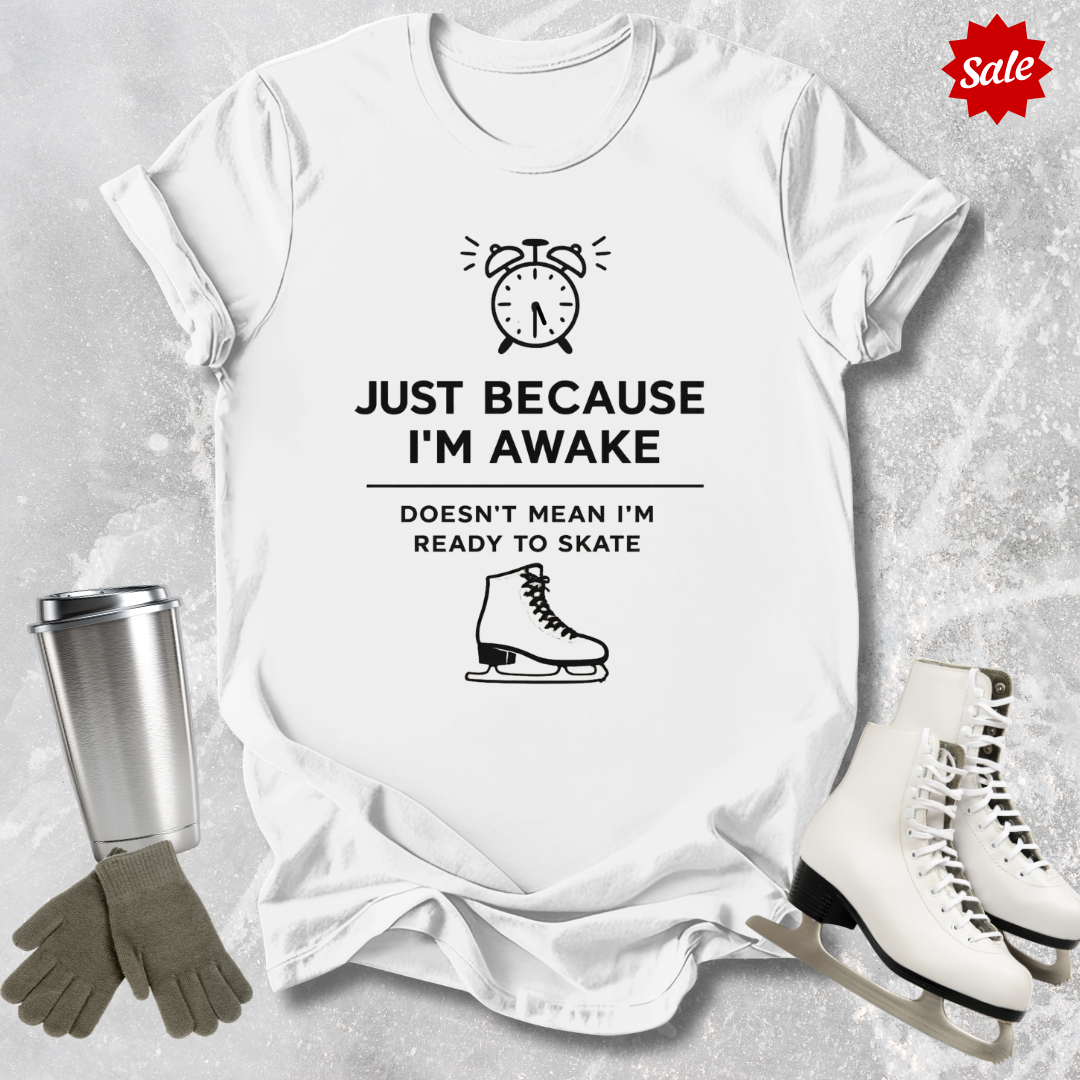 White t-shirt with humorous graphic and  Just Because I'm awake doesn't mean I'm ready to figure skate text, surrounded by ice skates, gloves, and a tumbler on a textured surface.