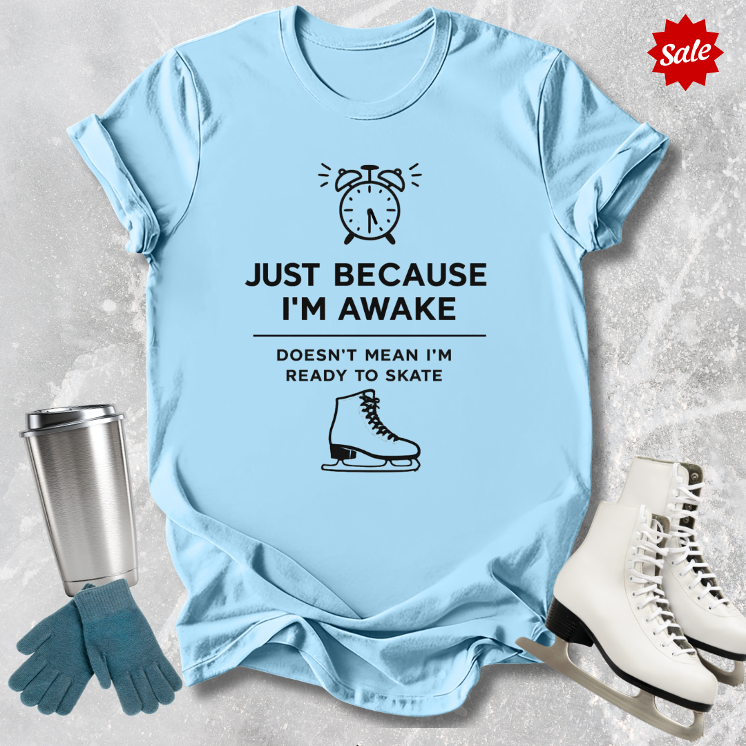 Light blue t-shirt with humorous graphic and  Just Because I'm awake doesn't mean I'm ready to figure skate text, placed on a textured surface with ice skates and gloves.