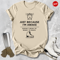 Beige t-shirt with humorous graphic and  Just Because I'm awake doesn't mean I'm ready to figure skate text, placed on a textured surface with ice skates and a tumbler for context.