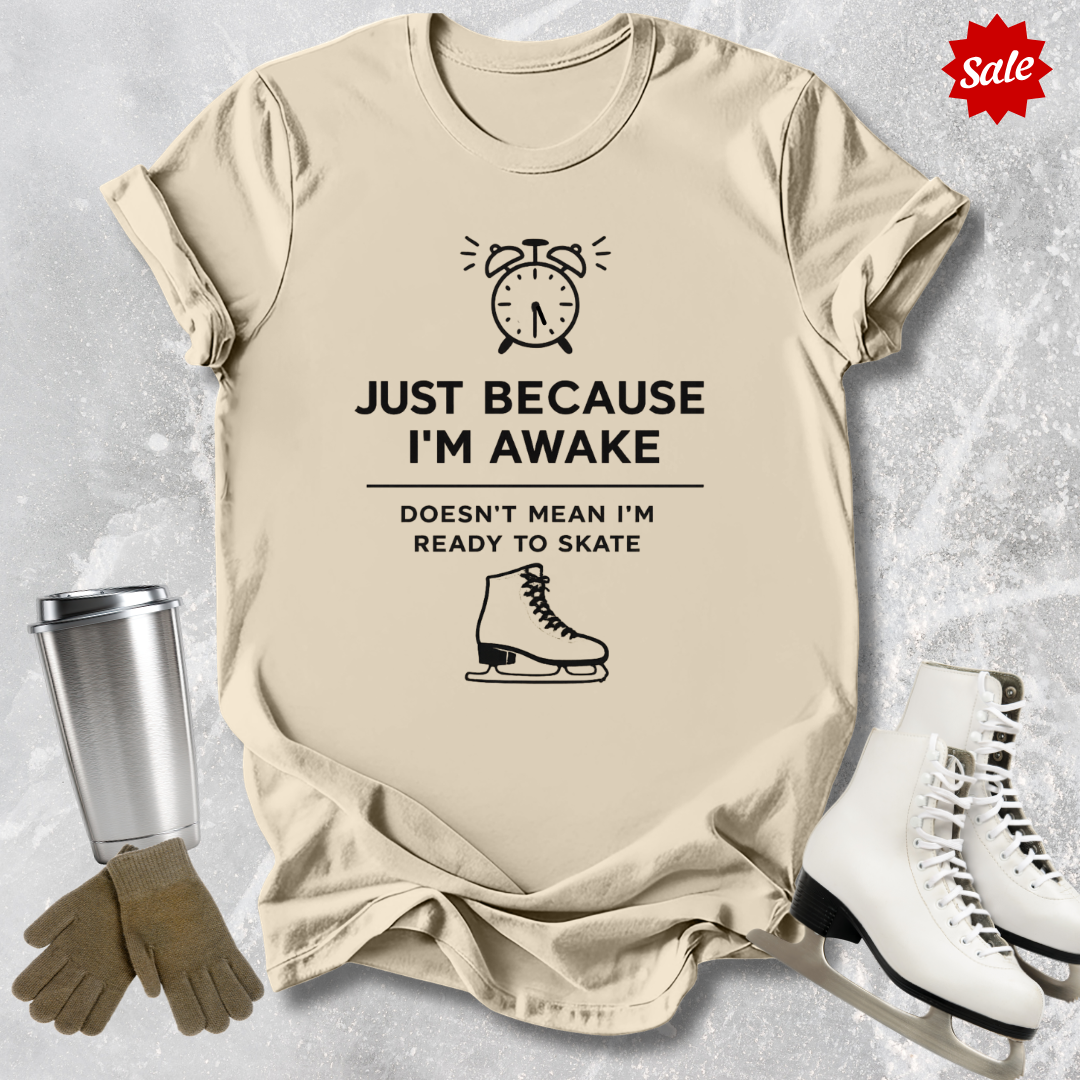 Beige t-shirt with humorous graphic and  Just Because I'm awake doesn't mean I'm ready to figure skate text, placed on a textured surface with ice skates and a tumbler for context.