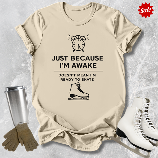 Beige t-shirt with humorous graphic and  Just Because I'm awake doesn't mean I'm ready to figure skate text, placed on a textured surface with ice skates and a tumbler for context.
