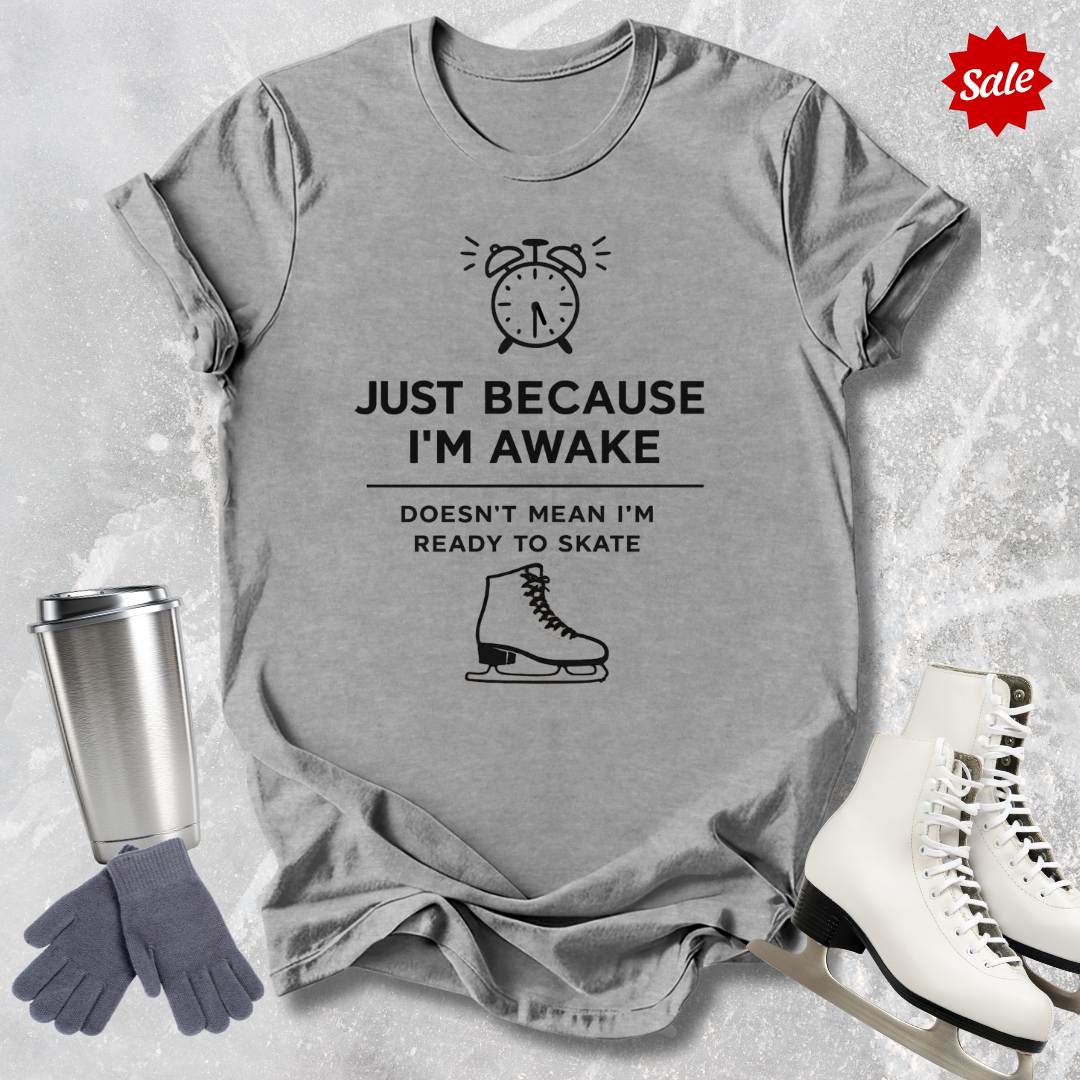 Gray t-shirt with humorous ice skating graphic and  Just Because I'm awake doesn't mean I'm ready to figure skate text, surrounded by ice skates, gloves, and a tumbler on a textured surface.