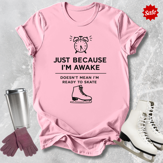 Pink t-shirt with humorous Just Because I'm awake doesn't mean I'm ready to figure skate text and graphics, placed on a textured surface with a cup, gloves, and skates for scale.