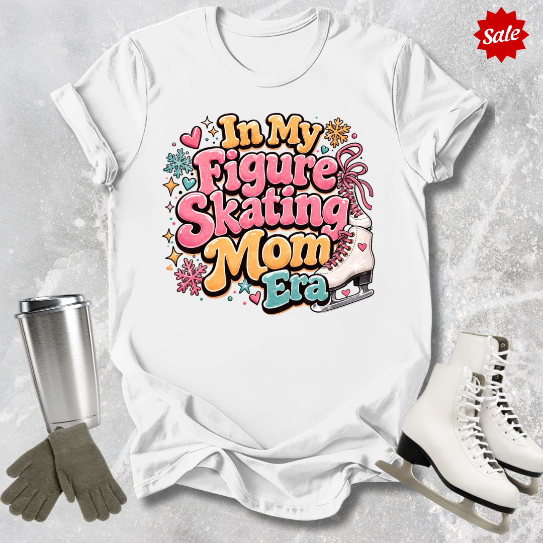 White t-shirt with 'In My Figure Skating Mom Era' design, surrounded by ice skates and a tumbler on a textured surface.