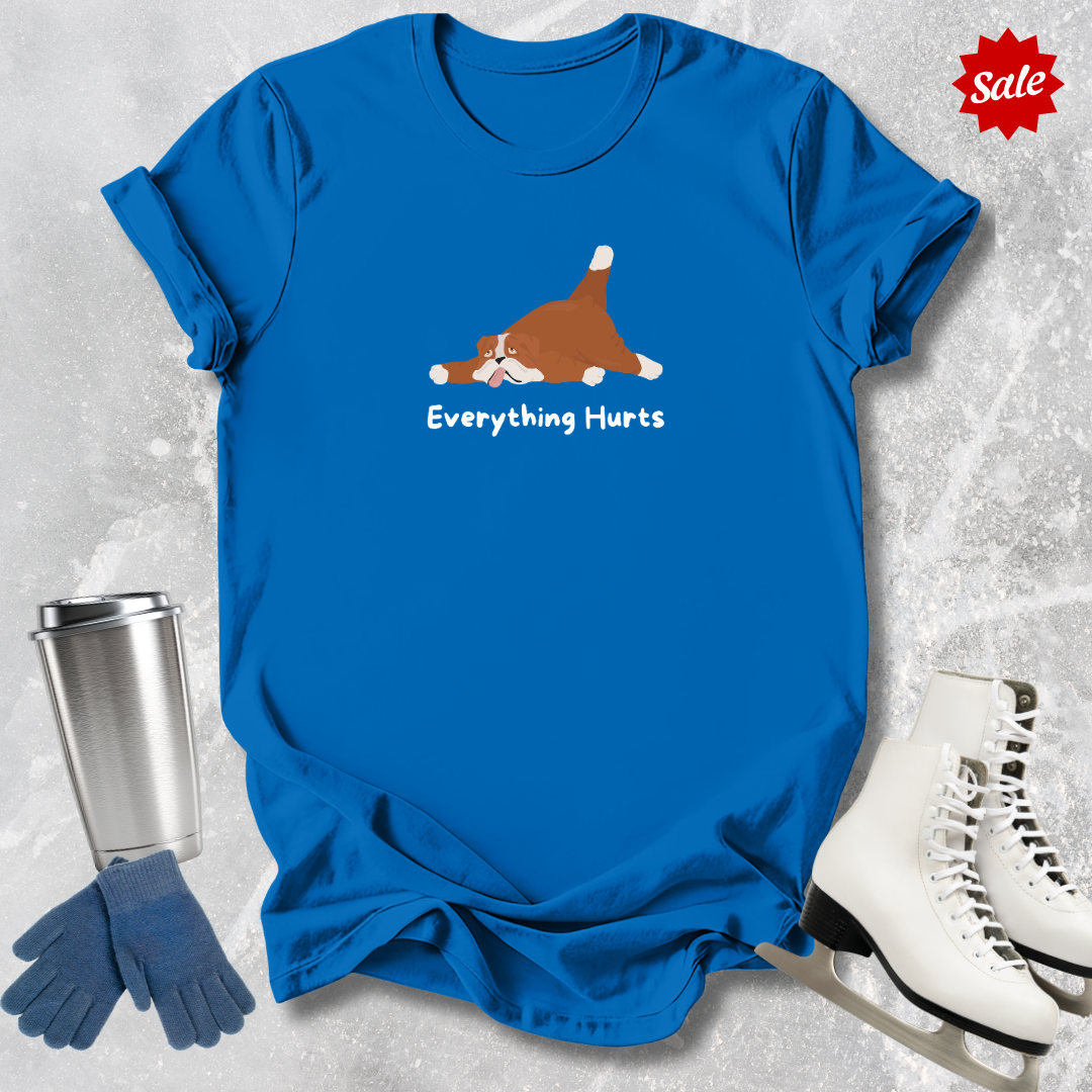 Blue t-shirt with a dog graphic and 'Everything Hurts' text, surrounded by a silver tumbler, blue gloves, and white ice skates on a textured gray background.