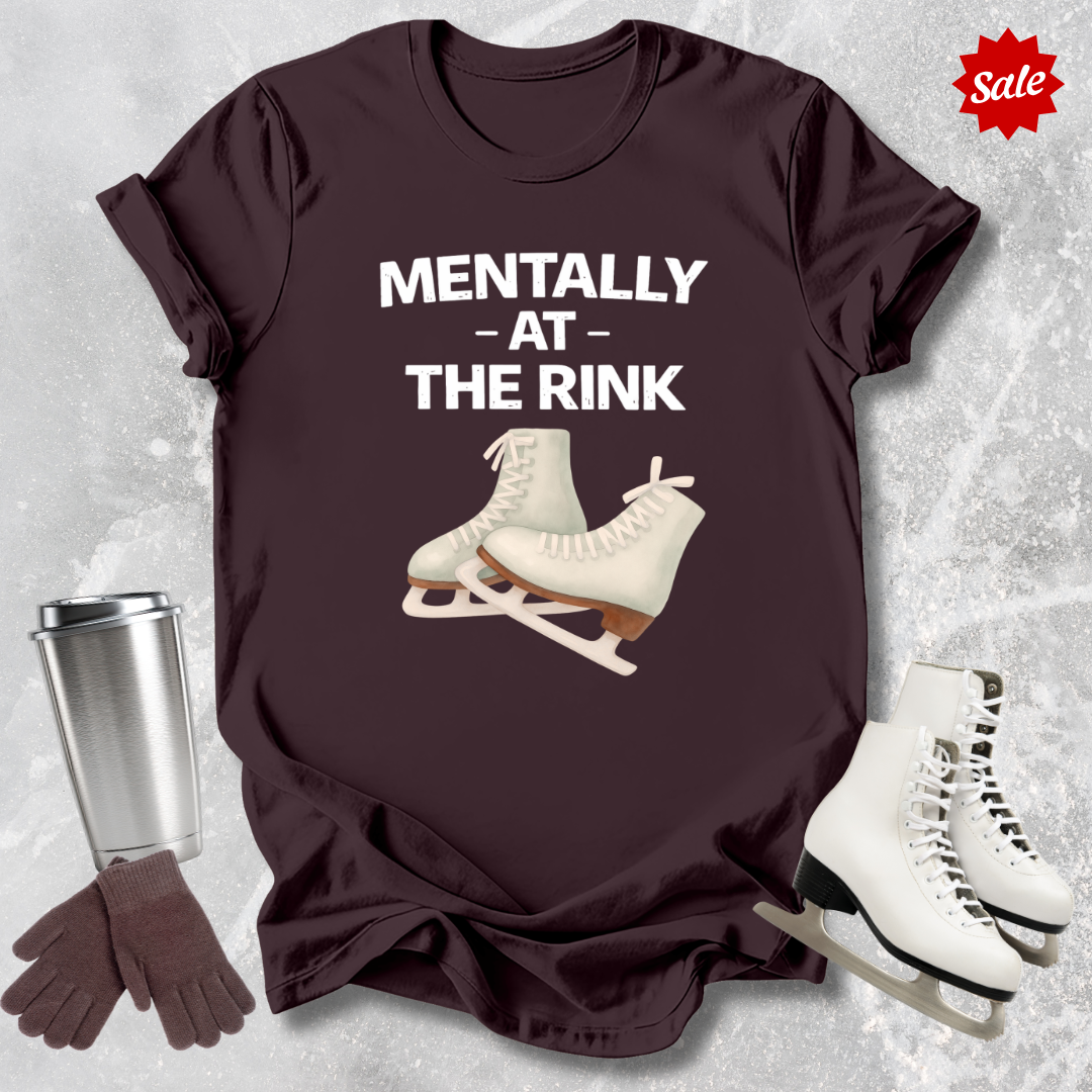 Maroon t-shirt with ice skates and text 'Mentally at the Rink' on a textured surface with accessories.