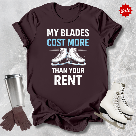 Brown t-shirt with 'MY BLADES COST MORE THAN YOUR RENT' text, next to a silver tumbler, black gloves, and white figure skates on a textured gray background.