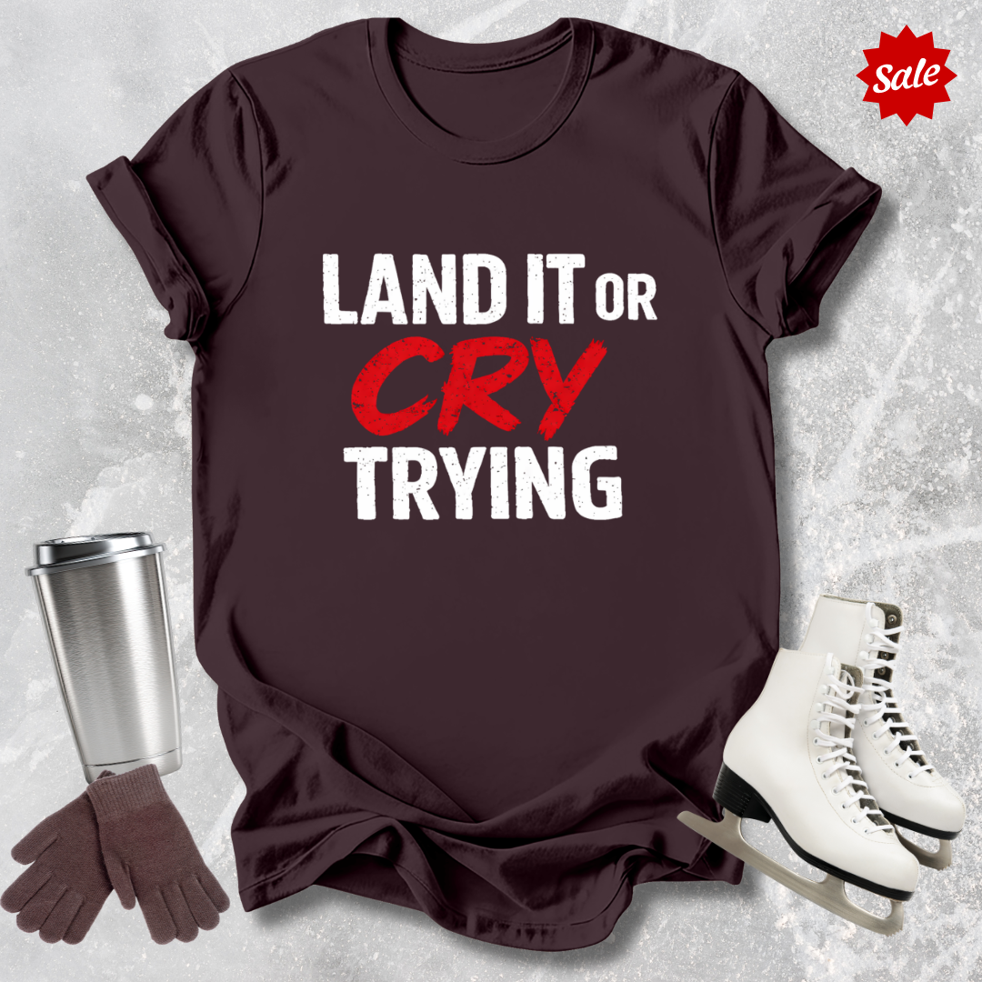 Brown  t-shirt with 'LAND IT OR CRY TRYING' text, next to a silver tumbler, black gloves, and white skates on a textured gray background.