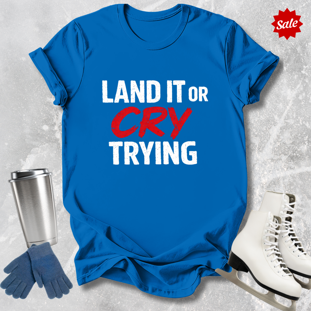 Blue t-shirt with 'LAND IT OR CRY TRYING' text, next to a silver tumbler, black gloves, and white skates on a textured gray background.